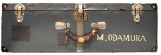 Vieille valise en cuir avec poignée ornée et attaches en métal. Le nom 'M.ODAMURA' est lisible près de la poignée.