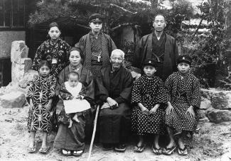 Black & white group family portrait in a garden. Everyone is dressed in traditional yukata. Three youths are wearing hats.