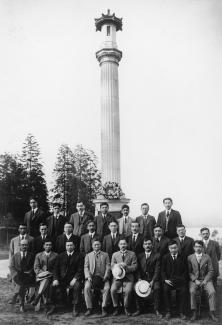 Black & white group portrait of well dressed men in front of an outdoor memorial. Some hold their hats. Trees in background.