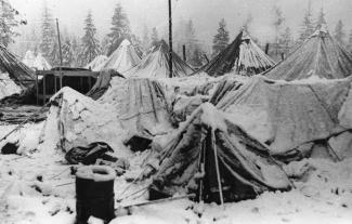 Black & white photo of group of tents covered in snow, some collapsed under weight of snow. Trees visible in background.
