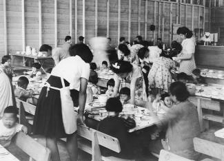 Black & white photo of women tending to children at meal time in a warehouse converted into a dining hall.