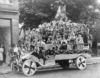 Black & white photo of parade float covered in Japanese and English flags carrying Japanese Canadian women in kimono.