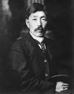 Black & white studio portrait of Japanese Canadian man with moustache wearing a suit with striped tie, holding hat.