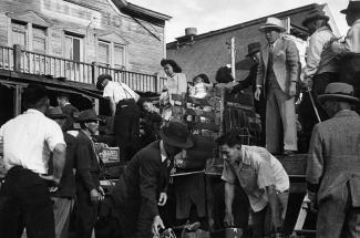 Black & white photo of men wearing suits and hats handling luggage around a truck with people standing on the truck bed.