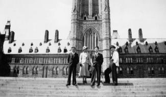 Three Japanese Canadian men and one woman stand for a portrait in front of the Centre Block of the Parliament Building.