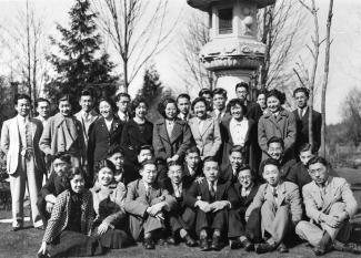 Black & white group photo of Japanese Canadian youth in coats and jackets. Leafless trees and evergreens in background.