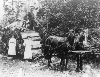 Black & white photo of man standing atop horse drawn log skid, two children sit next to him. Two women stand by the logs.