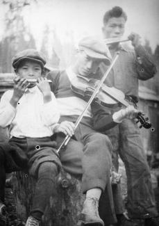 Black & white photo of three Japanese Canadian youths. Center figure playing violin with harmonica player at both sides.