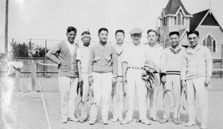 Black & white photo of eight Japanese Canadian men holding rackets at a tennis court with church in background.