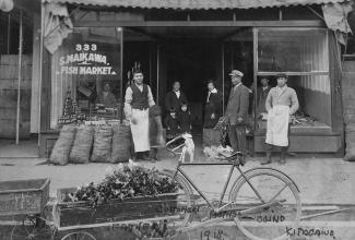 Black & white photo of Japanese Canadian shop clerks and patrons in front of fish market with bicycle and cart in foreground.