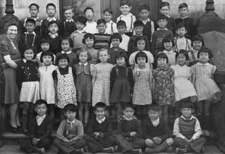 Black & white photo of multicultural group of young school children. Their teacher stands with them smiling.