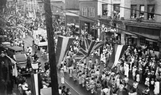 Black & white photo of Japanese Canadians parading in kimono down Powell Street with a Union Jack in the foreground.