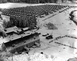 Black & white aerial photo of small shacks in rows at snowy compound. Trees separate huts from larger buildings and barn.