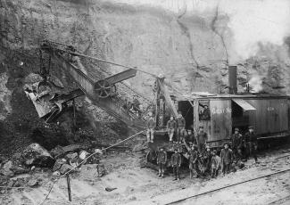 Black & white photo of a group of Japanese Canadian men standing in front of mining machinery for a group portrait.