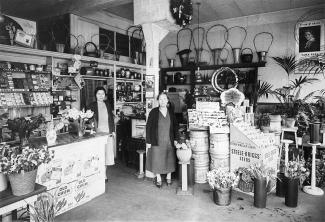 Black & white portrait of two Japanese Canadian women in a florist’s shop. Plants and flowers in buckets surround them.