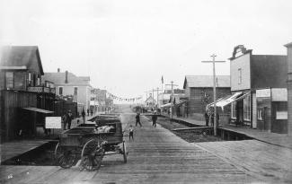 Black & white photo of boardwalk street lined by elevated wooden buildings. Wagon in foreground, men and child in distance.