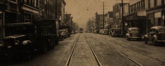Historical early west coast streetscape photo in sepia. Tram tracks in the centre with cars parked on either side. 