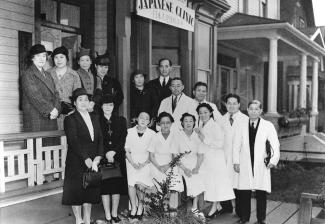 Black & white portrait of hospital staff on Japanese Clinic porch. Half in medical uniform, half in formal dress.