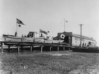 Black & white photo of boats resting on elevated wood platform. Japan flags raised among boats and warehouse in background. 