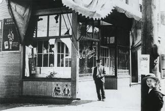 Black & white photo of man standing in front of broken store windows. Boy in hat stands in the foreground.