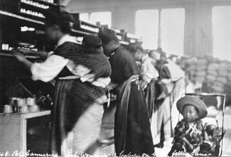 Black & white photo of row of Japanese Canadian women at work, two with babies on their backs, a toddler sits behind them.