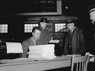 Image en noir et blanc d’un Canadien japonais, tête basse, avec deux officiers et un homme assis avec machine à écrire.