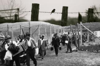 Black & white photo of Japanese Canadian men carrying their belongings into camp. Wooden building frames on either side.