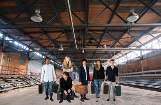 Montage of colour photos of youth holding suitcases, imposed over interior photo of warehouse with rafters at each side.