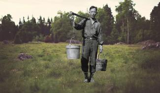 Black and white photo of Japanese Canadian man carrying buckets. Image of grassy field with forest in background.