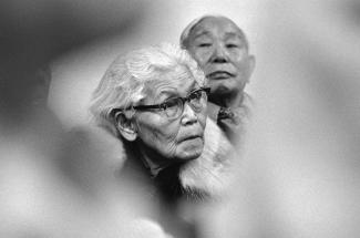 Black & white photo with blurred vignette foreground and two Japanese Canadian elders at center.