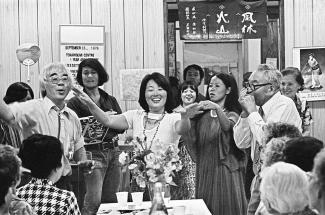 Black & white photo of Japanese Canadians gathered around a smiling woman stretching out her hands addressing the group.