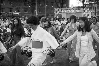 Image en noir et blanc de jeunes accompagnés d’aînés en tenues traditionnelles japonaises dansant dehors, main dans la main.