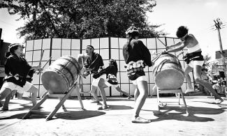 Black & white photo of taiko drumming group performing outdoors with large shoji screen in background.
