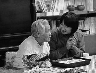 Black & white photo of an elder Japanese Canadian man talking with a young Japanese Canadian man at a table with food.