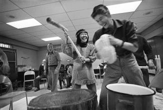Image en noir et blanc de deux jeunes et un aîné dans des vêtements des années 70. Ils rient en pilant du riz en mochi.