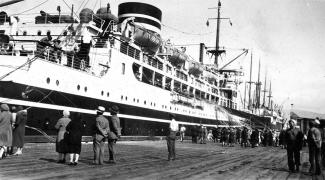 Black & white photo of large boat harboured at dock, with some passengers aboard and several people standing on the dock.