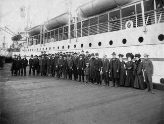Black & white photo of a row of Japanese Canadians standing on a pier in front of a large steamship with four lifeboats.