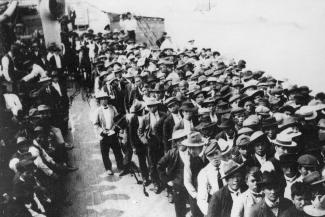 Black & white photo of crowd of Japanese men wearing suits and hats, mostly standing in lines with some seated to one side.