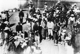 Black & white photo of many Japanese men crowded at a harbour, mostly wearing hats.