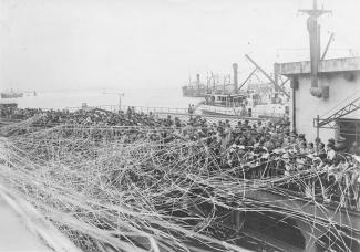 Black & white photo of a packed crowd waving streamers on a docked boat with water and other boats in the background.