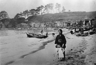 Black & white photo of woman and child in traditional Japanese dress standing on beach with village and hills in background.